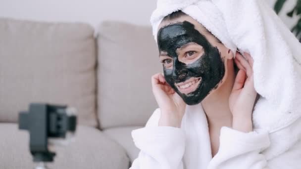 Woman in a white robe is applying black face mask and is being filmed — Stock Video