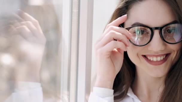 Hermosa mujer en gafas está cerca de la ventana sonriendo — Vídeos de Stock