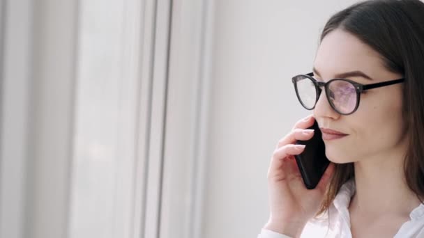 Mujer bonita en gafas está haciendo una llamada cerca de la ventana y sonriendo — Vídeos de Stock
