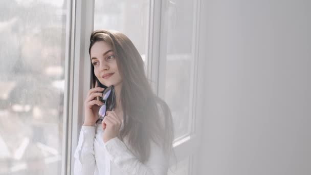 Mooie vrouw met bril belt bij het raam en glimlacht. — Stockvideo