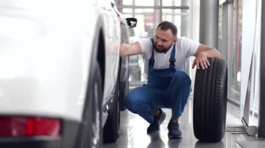 Mavi üniformalı tamirci arabanın tekerleğini içeride değiştiriyor.