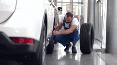 Mavi üniformalı tamirci arabanın tekerleğini içeride değiştiriyor.