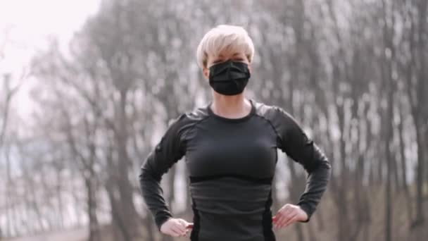 Een vrouw van middelbare leeftijd met een masker doet ochtendtraining buiten. — Stockvideo