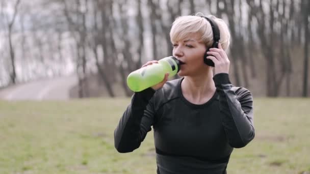 Mujer atlética en una máscara está escuchando música en los auriculares y el agua potable — Vídeo de stock