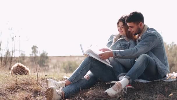 Jovem atraente está abraçando sua namorada lendo um livro no campo — Vídeo de Stock