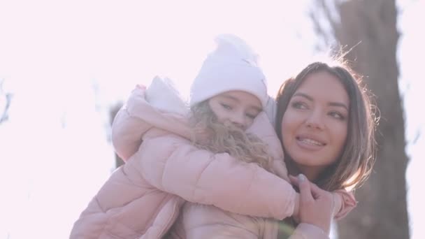 Die junge hübsche Frau trägt ihr süßes Mädchen auf dem Rücken beim Spazierengehen in einem Frühlingspark — Stockvideo