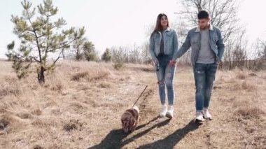 Kot giyen genç adam ve kadın buğday tarlasında bir köpekle yürüyorlar.