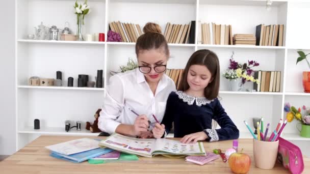 Мама в очках помогает брюнетке дочке делать домашнее задание дома — стоковое видео