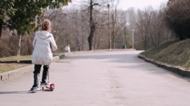 Küçük kız bir parkta scotter kullanıyor.