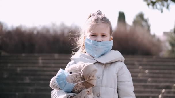 La petite fille dans un masque jetable tient un ours en peluche dans un masque à l'extérieur — Video