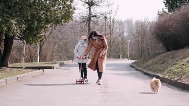 マスクを着たスクーターの母親と娘が犬と公園を歩いています — ストック動画