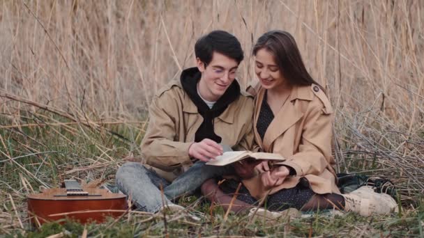 Pareja joven sentada en el campo y leyendo el libro en voz alta — Vídeos de Stock