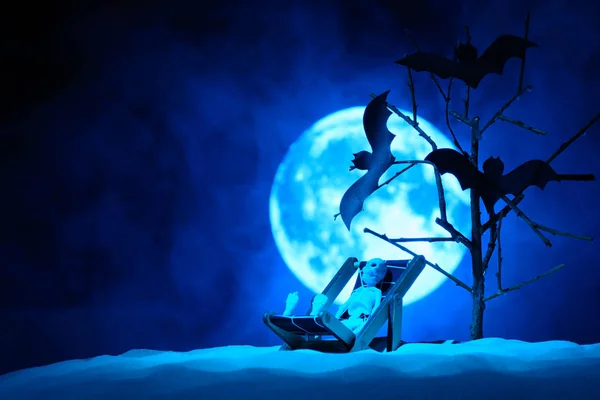 Un esqueleto de juguete en una silla para relajarse y tres murciélagos en un árbol, la luna llena en la niebla detrás — Foto de Stock