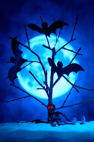 Esqueleto de brinquedo com olhos vermelhos e três morcegos em uma árvore, a lua cheia na névoa atrás — Fotografia de Stock
