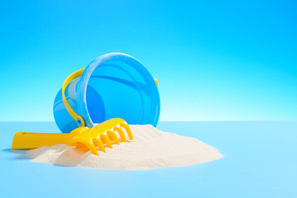 Toy plastic pail and a rake on the sand. Blue background with copy space