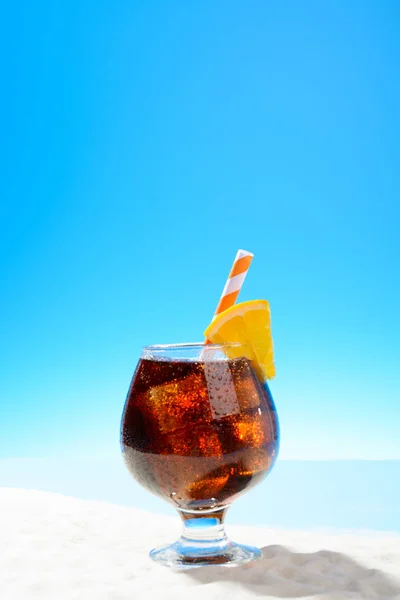 在沙质海岸的玻璃上用冰和一片橙汁提神可乐 — 图库照片