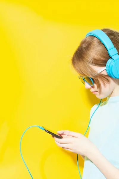Teenager s culíkem, nosit sluneční brýle a modrá sluchátka poslouchat hudbu s smartphone na žlutém pozadí — Stock fotografie