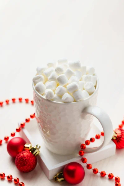 White mug with marshmallows with Christmas decorative balls on a white table. Festive composition, top view.