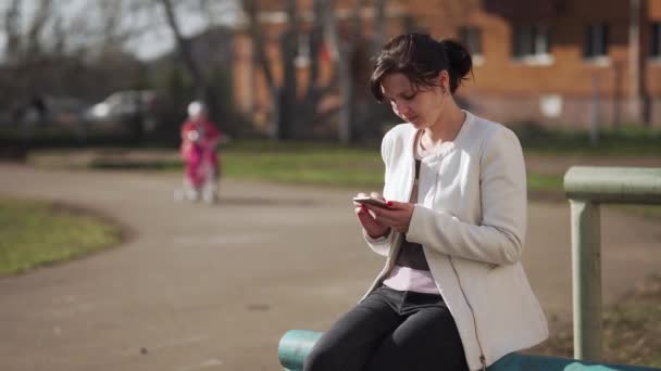 Genç bir kadın akıllı telefon kullanarak parkta oturur ve çocuğa bisikletin en üst parmağını gösterir.. — Stok video