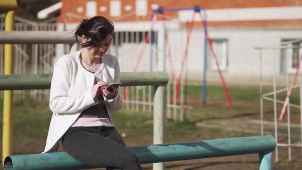 Retrato de una joven al aire libre usando un teléfono inteligente. Una chica con una chaqueta blanca se sienta en un parque y marca un mensaje en el teléfono . — Vídeos de Stock