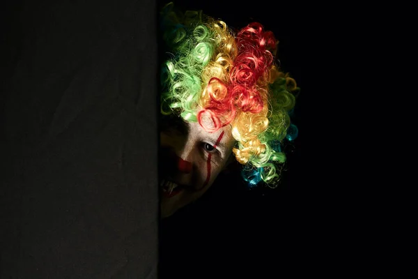 Close-up de um palhaço assustador espreitando ao virar da esquina de uma parede preta. Ele está usando uma peruca colorida e presas afiadas . — Fotografia de Stock