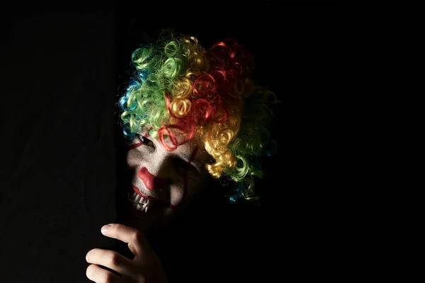 Крупним планом страшний клоун підглядає за рогом чорної стіни. Він одягнений в кольорову перуку і гострі ікла . — стокове фото