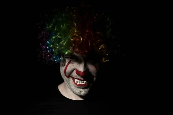 Close-up of a creepy clown peeping out of the darkness. He is wearing a colored wig and sharp fangs. — Stock Photo, Image