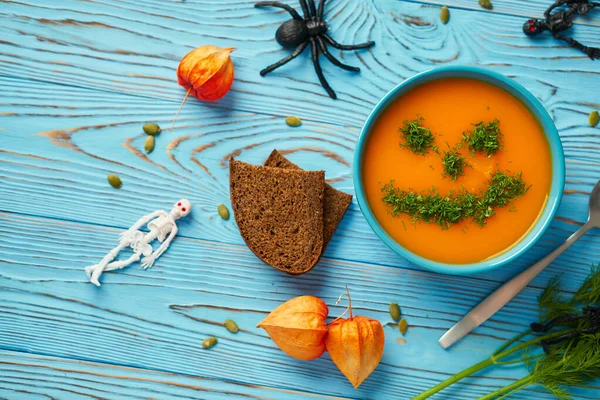 Autumn cream soup garnished with dill in the form of funny faces Halloween pumpkins. Healthy pumpkin and carrot soup puree in a blue bowl on a wooden background. Halloween holiday concept, top view. — Stock Photo, Image
