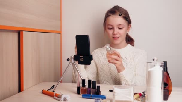 Linda chica joven maquillaje blogger muestra su nuevo perfume a los suscriptores. Graba vídeo para un blog en las redes sociales usando una cámara de teléfono inteligente en un trípode . — Vídeo de stock