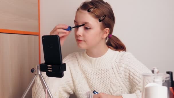 Linda joven blogger maquillaje pestañas rímel. graba vídeo para un blog en las redes sociales usando una cámara de teléfono inteligente en un trípode . — Vídeo de stock