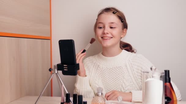 A cute young girl blogger on makeup demonstrates the technique of using a makeup brush. records video for a blog on social networks using a smartphone camera on a tripod. — Stock Video