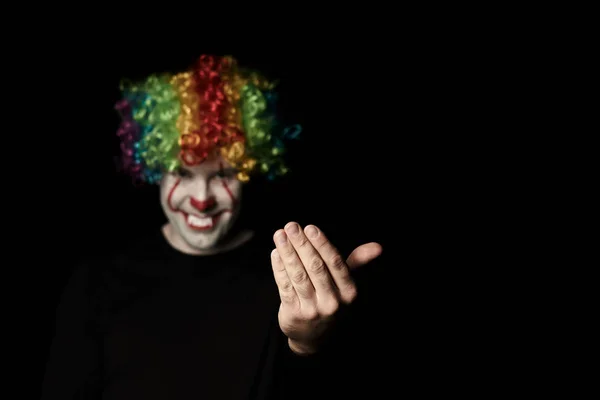 Ein gruseliger Clown mit farbiger Perücke ruft mit einer Handgeste nach sich. steht in einem dunklen Raum und lächelt. — Stockfoto