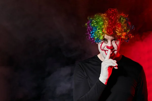 Creepy scary clown in a colored wig. Shows a gesture of silence with a finger at the lips. Empty space for text on the left. — Stock Photo, Image