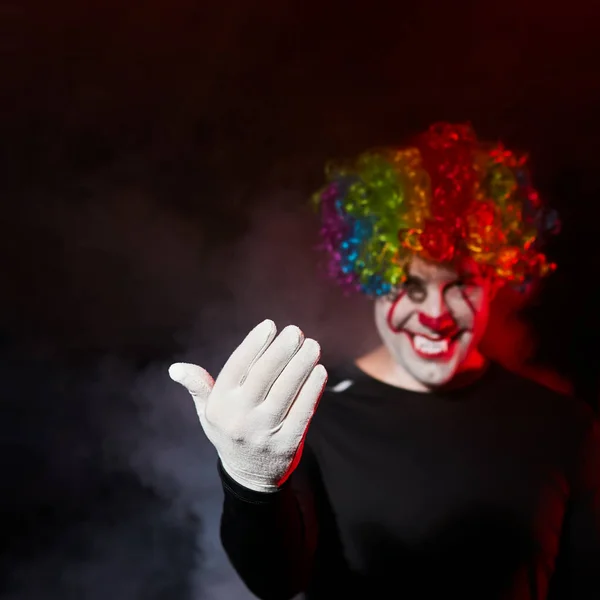 Ein gruseliger Clown mit farbiger Perücke lächelt und winkt sich mit einer Handbewegung zu. steht in einem verrauchten dunklen Raum. — Stockfoto