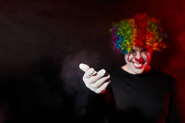 Un clown inquietante con una parrucca colorata sorride e si fa cenno con un gesto della mano. Sta in una stanza buia e fumosa . — Foto Stock