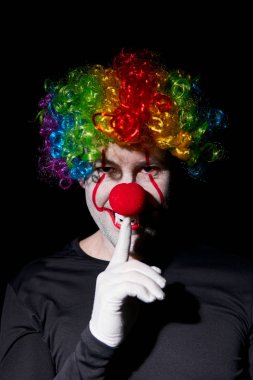 Renkli peruklu ürkütücü palyaço. Dudaklarında bir parmak ile sessizlik jesti gösterir.