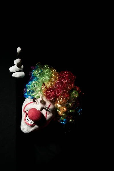 Enge clown gluurt om de hoek van een zwarte muur. Hij draagt een gekleurde pruik en scherpe hoektanden.. — Stockfoto