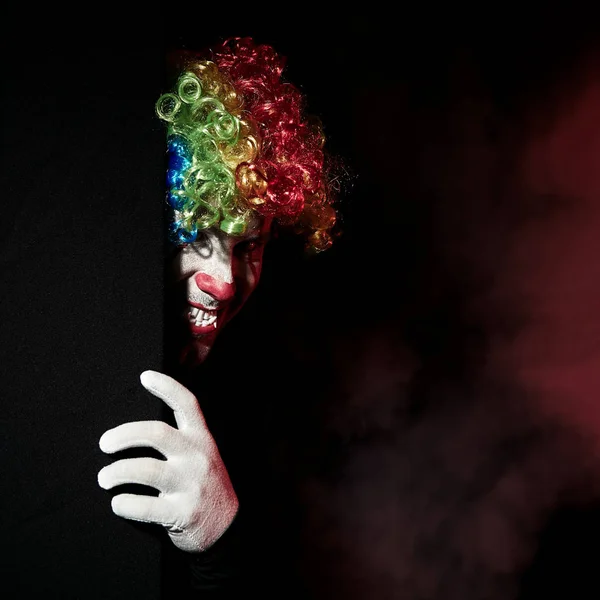 Страшний клоун підглядає за рогом чорної стіни. Він одягнений в кольорову перуку і гострі ікла . — стокове фото
