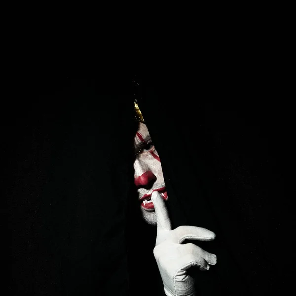 Ужасный страшный клоун в цветном парике выглядывает из-за черных занавесок. Показывает жест молчания с пальцем на губах . — стоковое фото