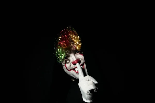 Zpoza černých záclon vykukuje děsivý klaun v barevné paruce. Ukazuje gesto ticha s prstem na rtech. — Stock fotografie