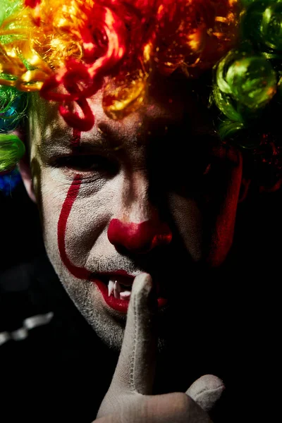 Pauroso clown spaventoso con una parrucca colorata. Mostra un gesto di silenzio con un dito sulle labbra . — Foto Stock