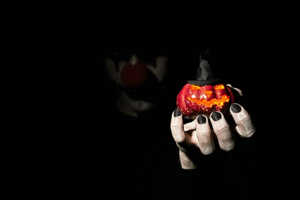 Жахливий жахливий клоун стоїть у темній кімнаті і на витягнутій руці тримає гарбуз на Хеллоуїн . — стокове фото