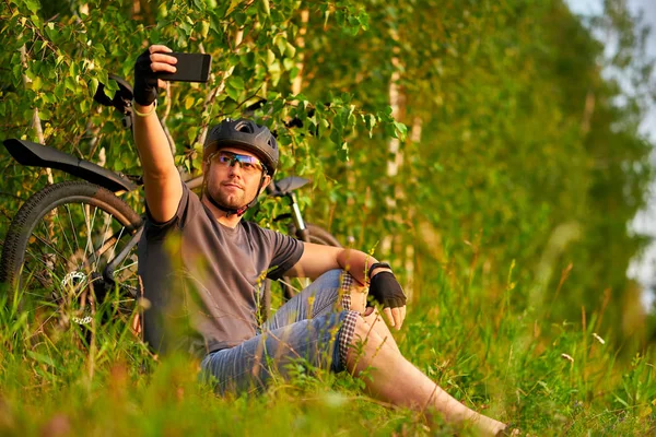 Samiec rowerzysta w kasku siedzi na trawie obok roweru, trzyma smartfona, robi selfie. — Zdjęcie stockowe