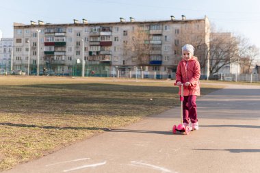 Pembe ceketli şirin kız okul bahçesinde scooter sürüyor. Sonbahar güneşli hava.