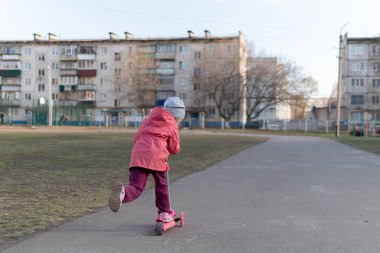Pembe ceketli şirin kız okul bahçesinde scooter sürüyor. Sonbahar güneşli hava.