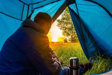Mavi ceketli genç bir adam çadırda oturur ve güneşin doğuşunu izler..