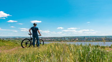 Bir erkek bisikletçi bir tepede duruyor ve şehre bakıyor. Kasklı ve bisikletli bir adam. Sırtı kameraya dönük..