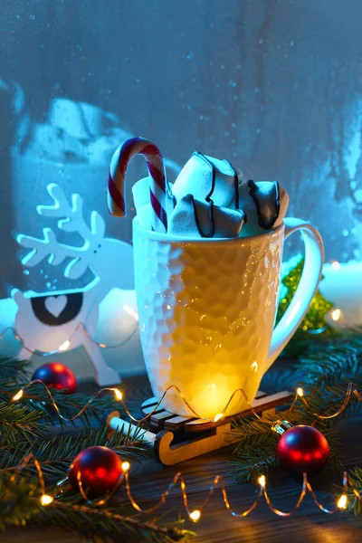 Белая кружка с горячим шоколадом и зефиром с рождественскими украшениями на подоконнике в зимний вечер у синего окна . — стоковое фото