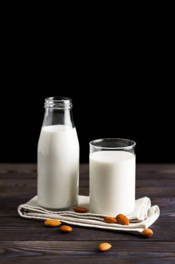 Bardakta badem sütü ve yeniden doldurulabilir cam şişe. Ahşap masa ve siyah arka planda badem fıstığı var..