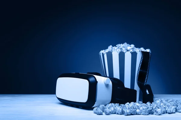 Composition with cinema film, a bucket of popcorn and VR headset on the wooden table. Toned in trendy classic blue color year 2020